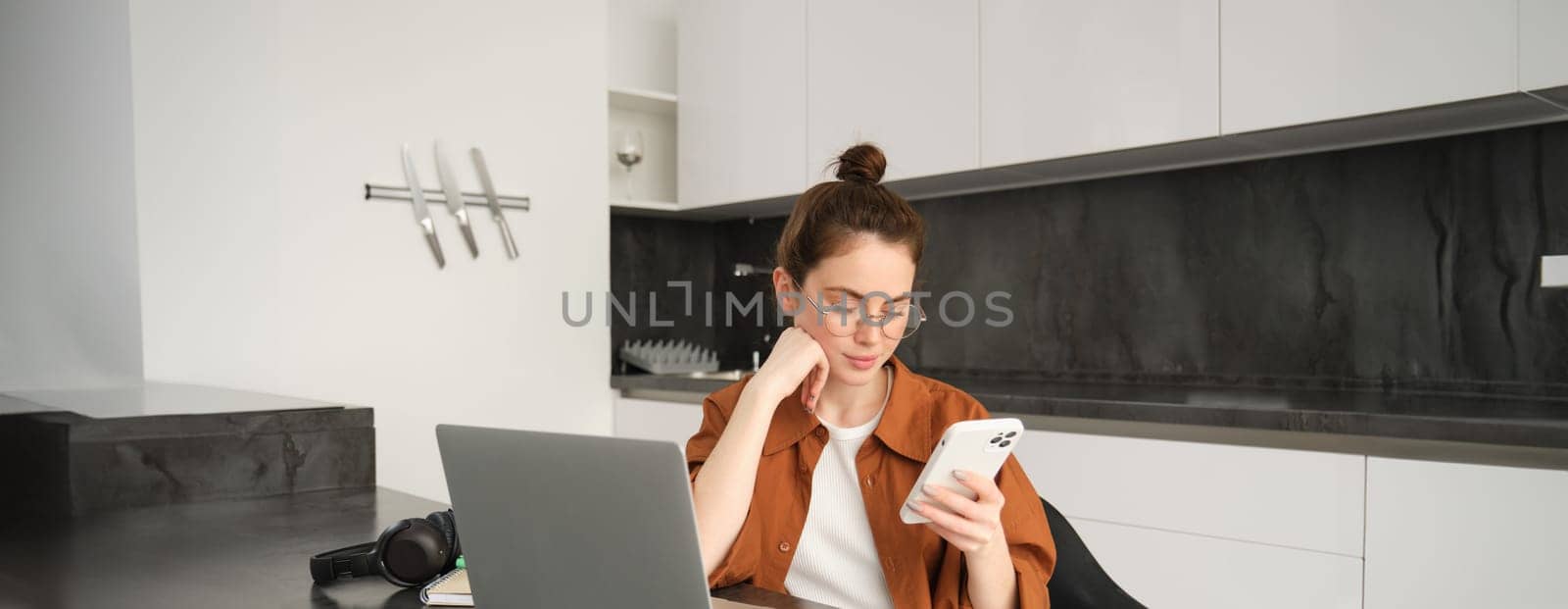 Portrait of young self-employed woman working on start up from home, freelancer programmer with computer, doing her task on computer, checking her mobile phone, reading message on smartphone.