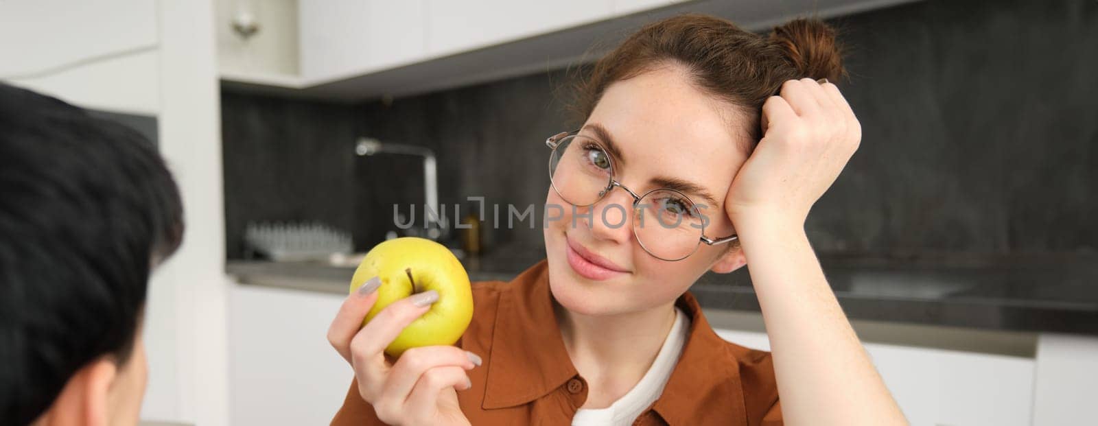 Close-up portrait of brunette woman at home, wearing glasses, eating apple in the kitchen and smiling, biting fruit by Benzoix