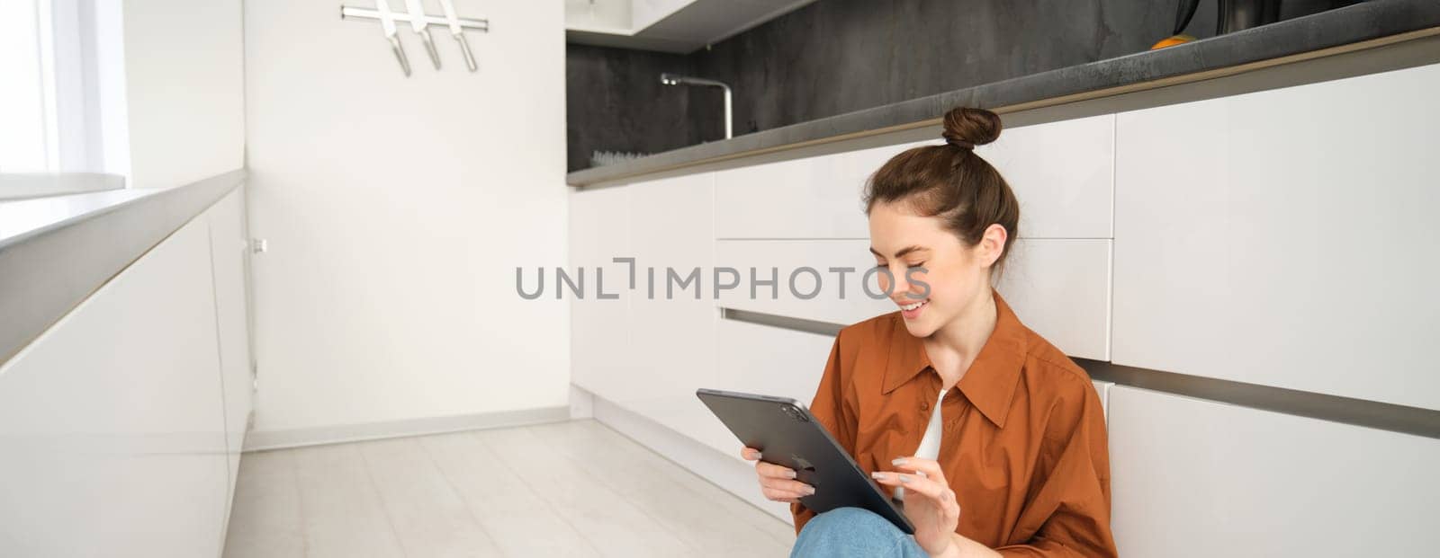 Portrait of pretty young woman at home, sitting on kitchen floor, looking at digital tablet, using gadget, reading on device by Benzoix