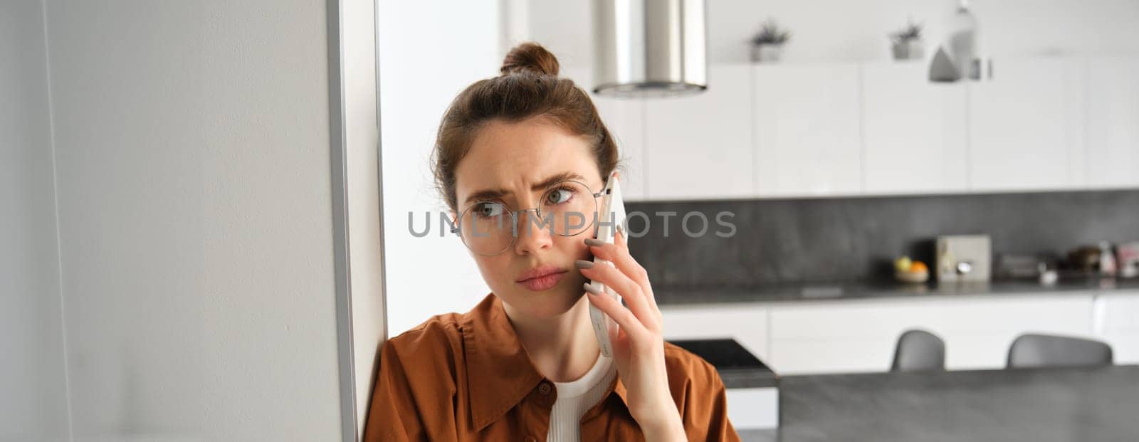 Portrait of woman with disappointed face, standing at home, answers phone call with concerned face, has difficult telephone conversation.