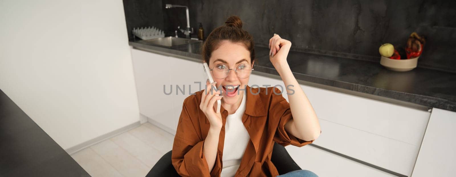 Excited young woman talking on mobile phone in front of laptop, sitting in kitchen with happy face expression, having a conversation.