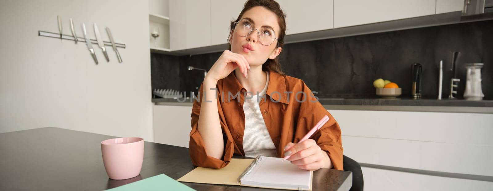 Portrait of woman thinking, sitting kitchen, holding pen, writing with thoughtful face, creative girl pondering over her project, making notes.