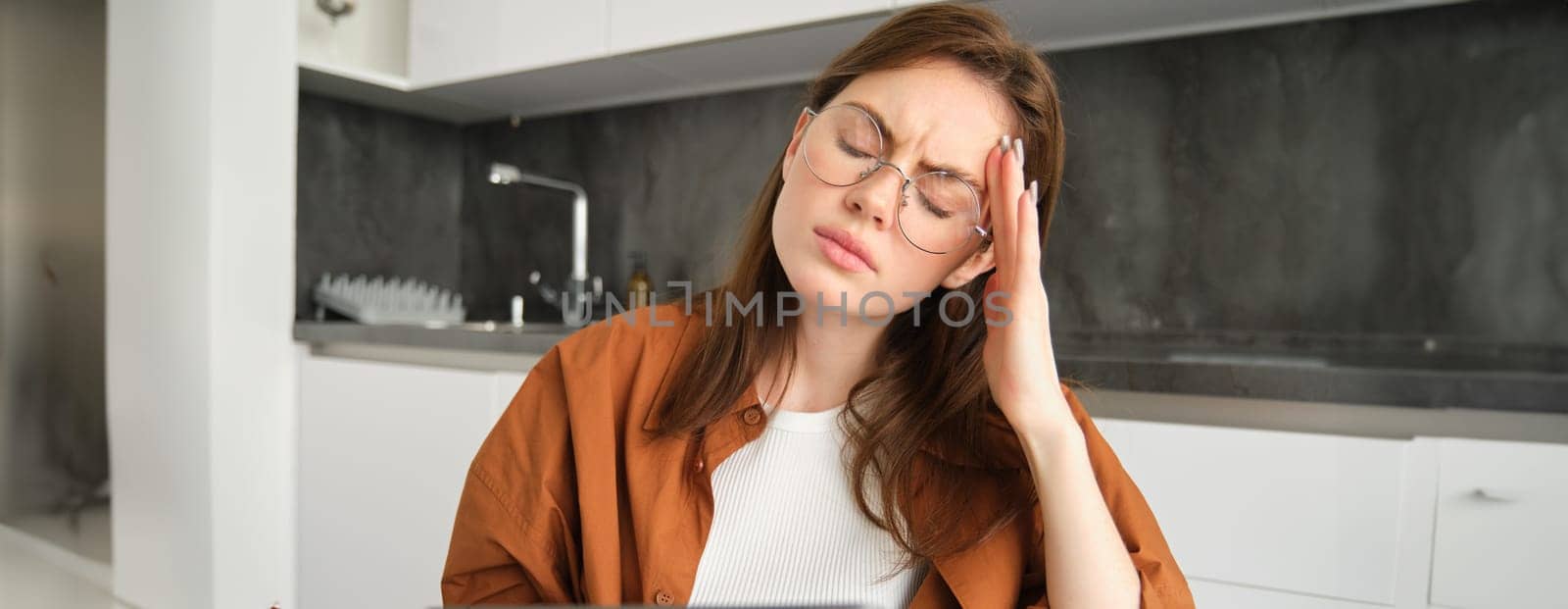 Portrait of young woman sitting at home with digital tablet, feeling unwell, touching head and frowning from pain, has headache or migraine, working while feeling sick.