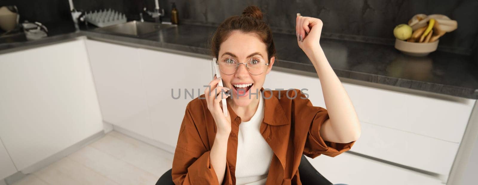 Portrait of woman receiving great news over the phone. Girl talking on mobile telephone and celebrating, laughing and making fist pump, dancing on her chair in kitchen by Benzoix