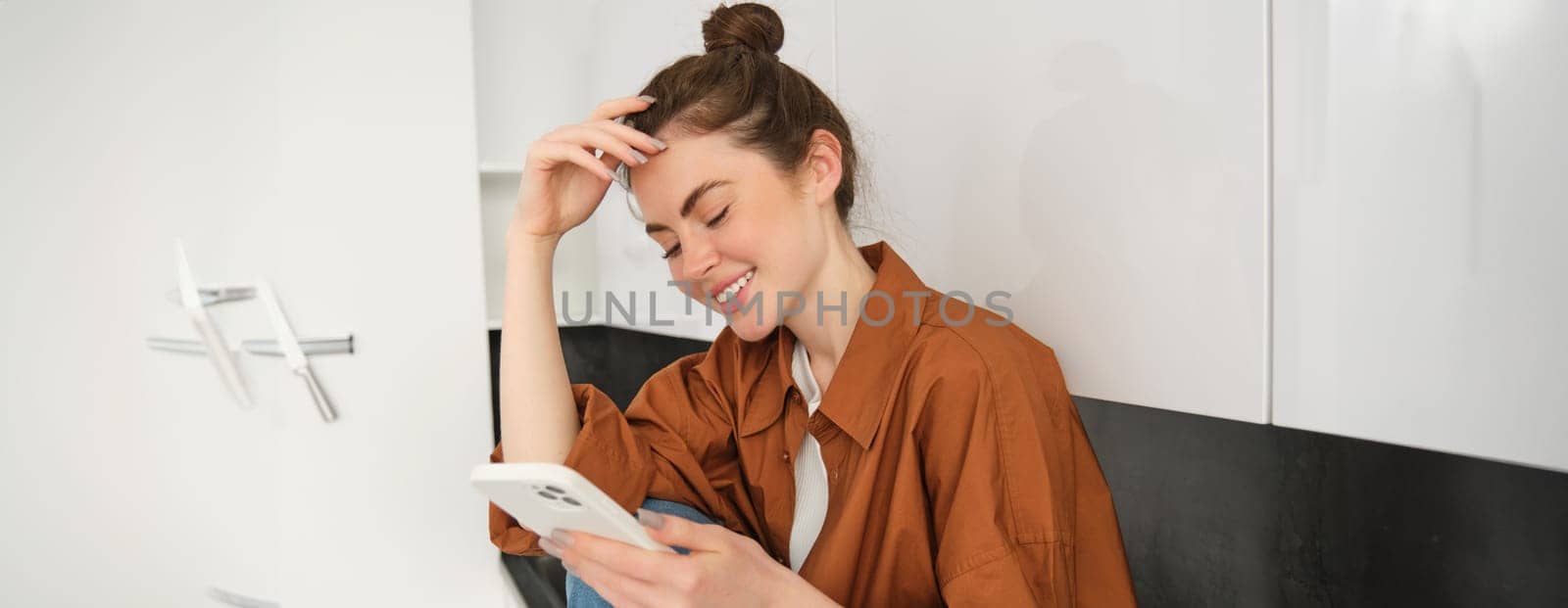 Young woman sitting at home on kitchen counter and using her smartphone. Concept of social media, leisure and mobile connection.