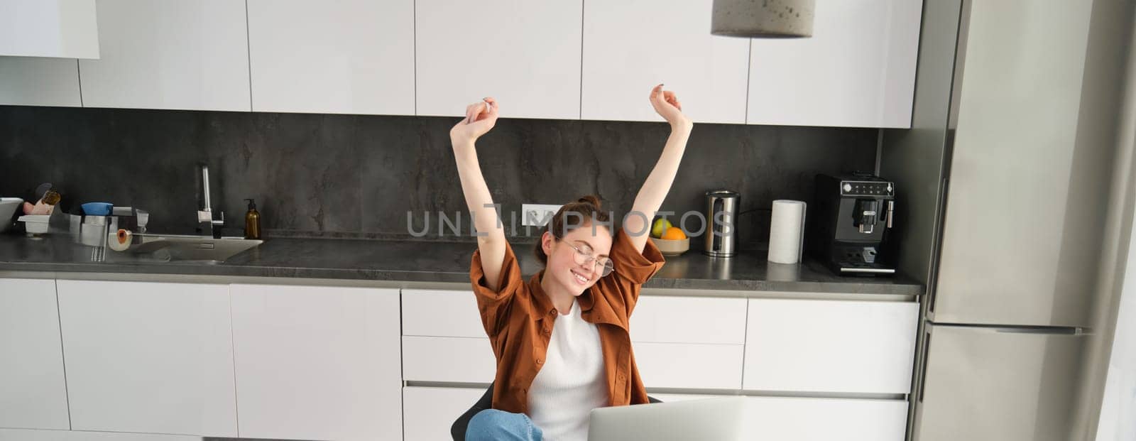 Productivity and lifestyle concept. Beautiful young woman finishes working, stretching hands with satisfied expression, sitting in kitchen with laptop and documents, has remote workplace at home.