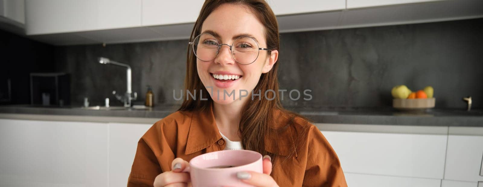 Close up portrait of beautiful brunette woman in glasses, resting, sitting in kitchen with cup of tea, holding mug, laughing and smiling.