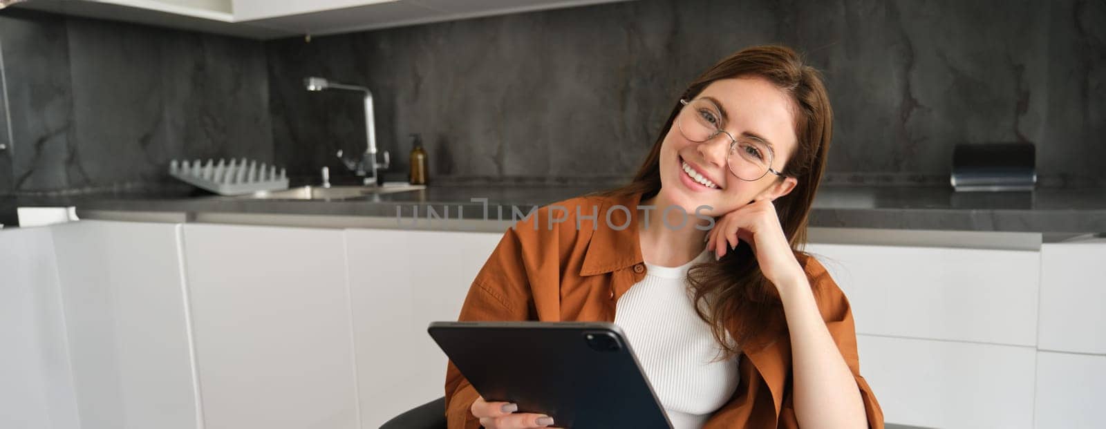 Portrait of beautiful smiling girl, wearing glasses, sitting in kitchen with digital tablet, working on remote from home.