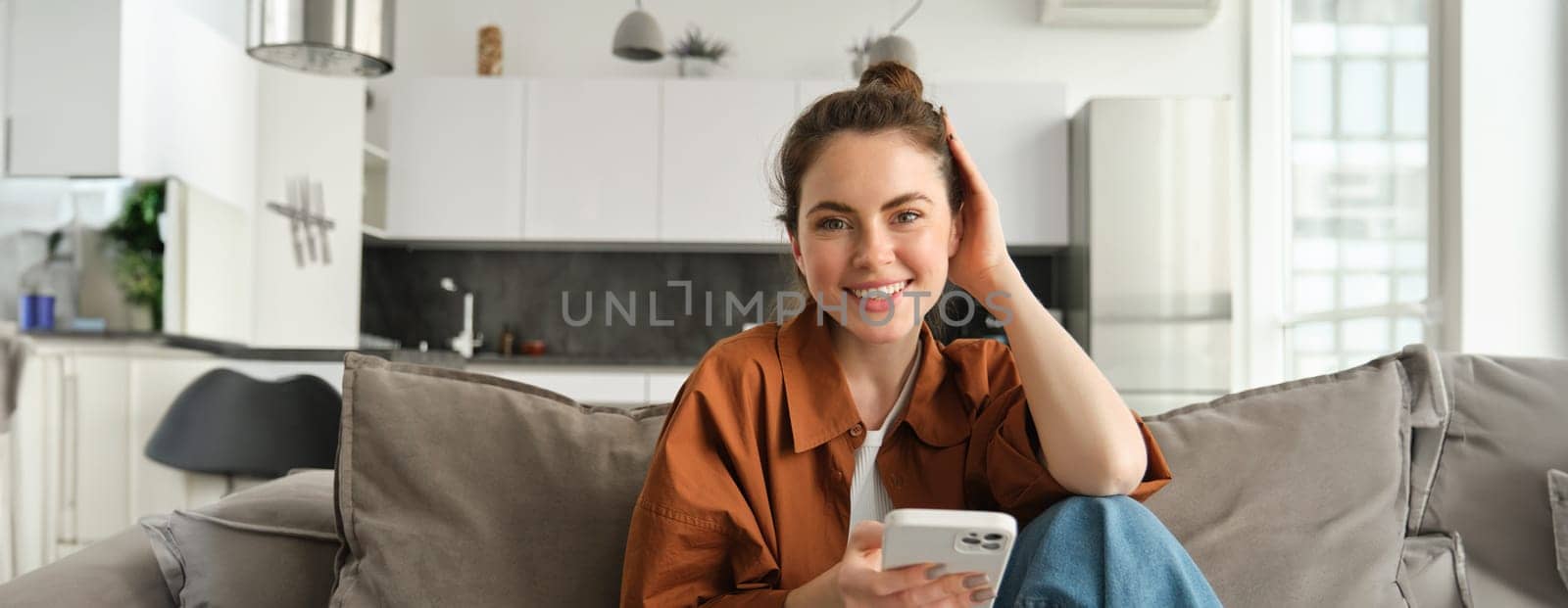 Beautiful smiling female model sitting on couch with smartphone, looking happy at camera, scrolling social media, messaging someone, doing online shopping.