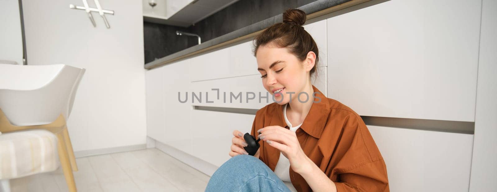 Portrait of young smiling woman, girl puts on her black wireless headphones, listens to music in earphones, sits on kitchen floor at home.