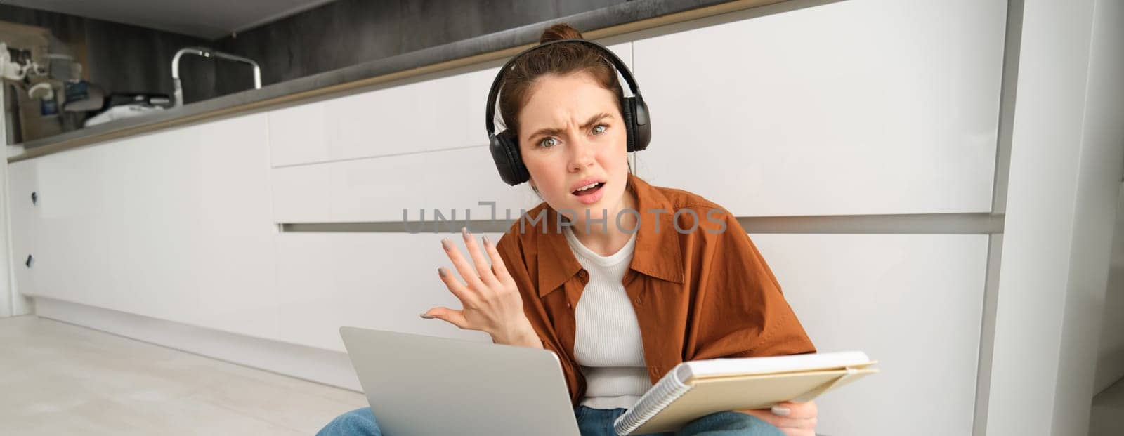 Portrait of annoyed and confused woman in headphones, sits on floor with laptop and notebook, looks frustrated, has issues in doing homework or studying.