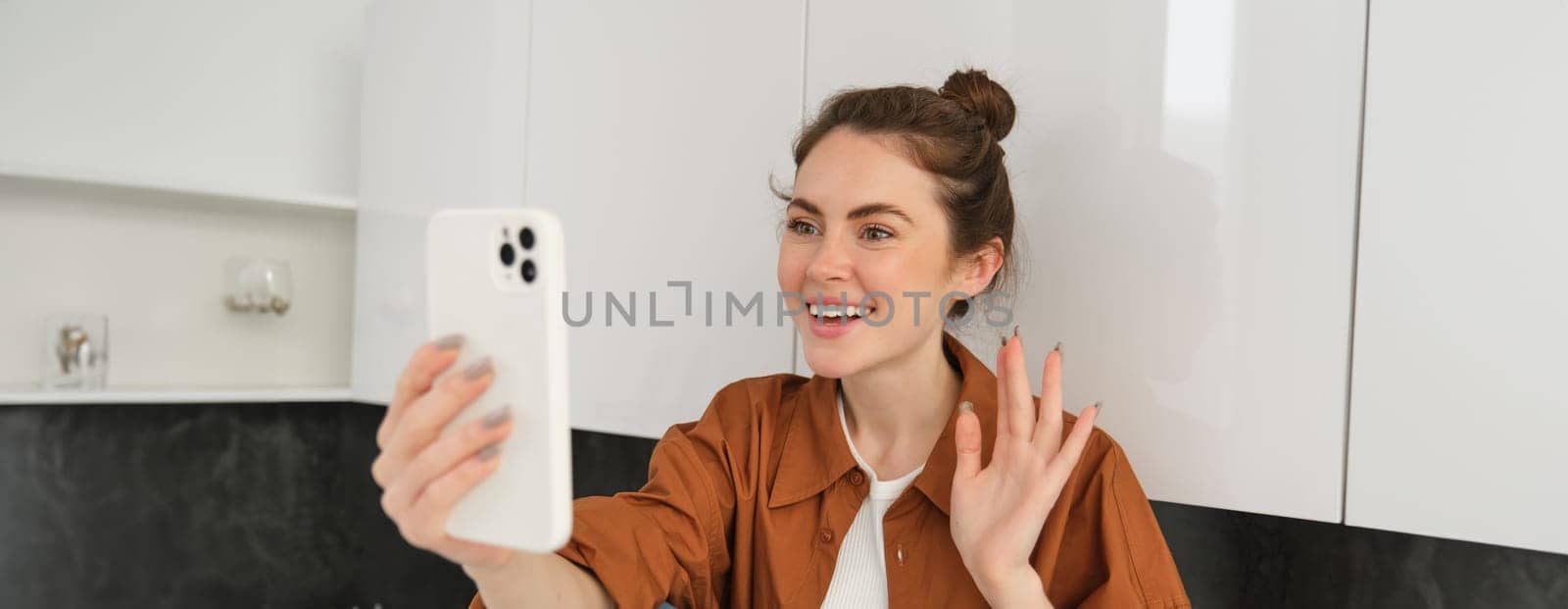 Portrait of young cheerful woman laughing and smiling during phone call, video chats with friend, sits on kitchen counter and talks to someone using smartphone app.