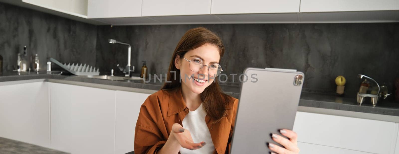 Portrait of young woman joins online course on digital tablet, having conversation, chatting on remote, sitting in kitchen.