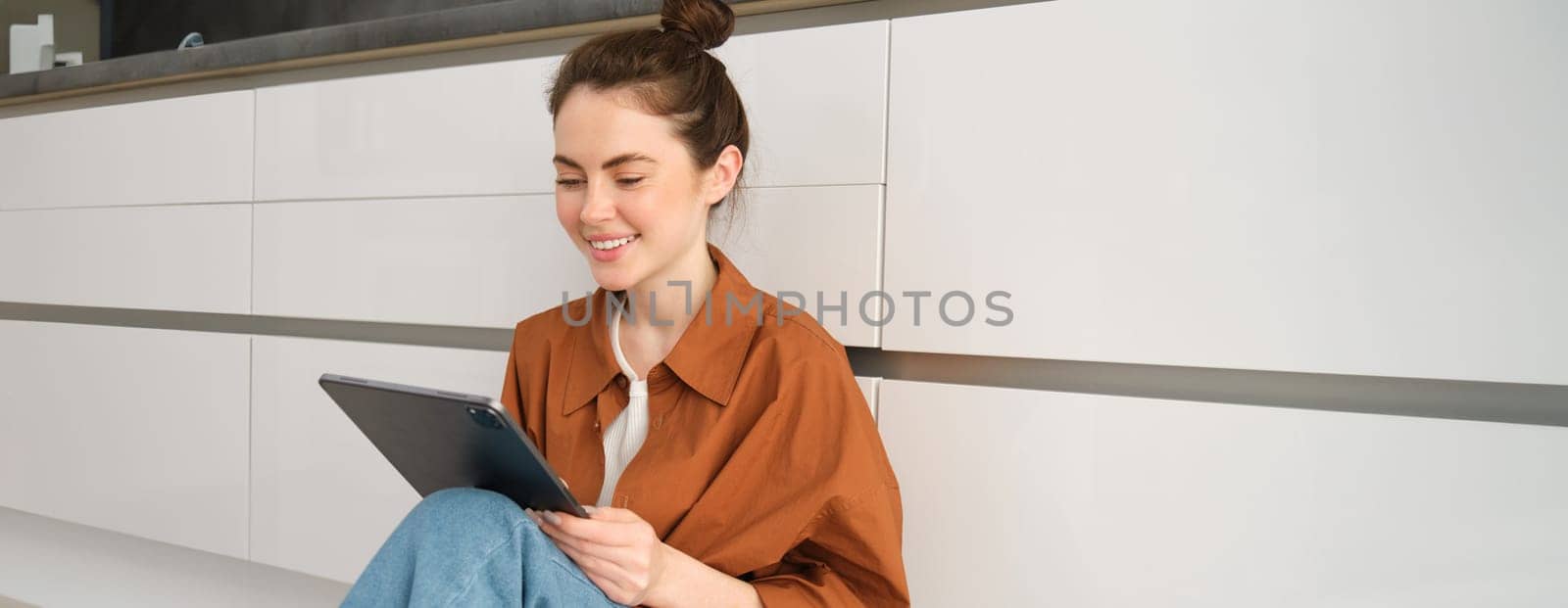 Portrait of young beautiful woman sitting on kitchen floor with digital tablet, browsing news feed, social media app on gadget, smiling and looking happy by Benzoix