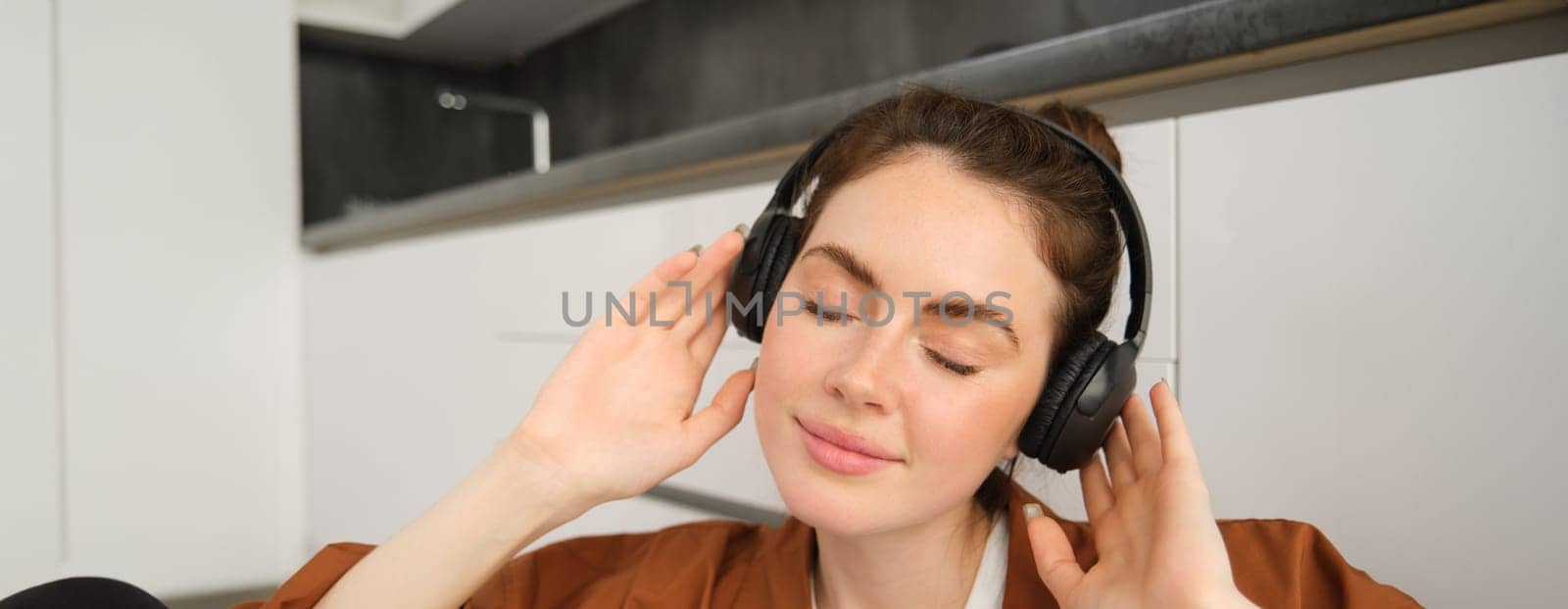 Close up shot of cute young woman in wireless headphones, listens to music and smiles, relaxes at home with favourite songs playing on her playlist by Benzoix