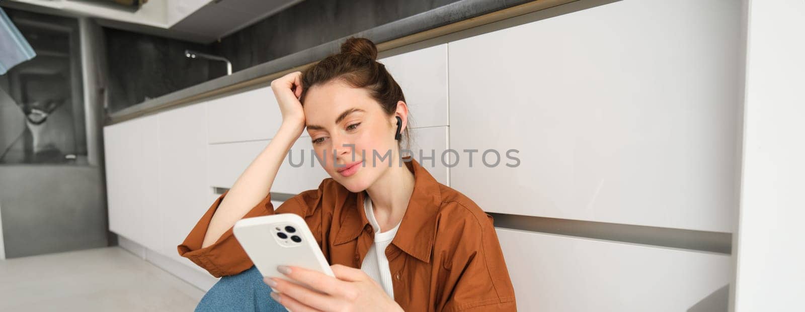 Portrait of beautiful smiling woman sitting on kitchen floor, scrolling social media on mobile phone, listening music in wireless headphones.