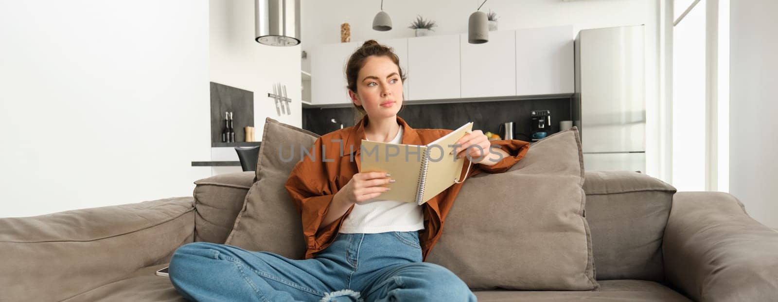 Young woman sits at home on sofa, reading her notes, holding notebook, studying for exam, student revising in her living room by Benzoix