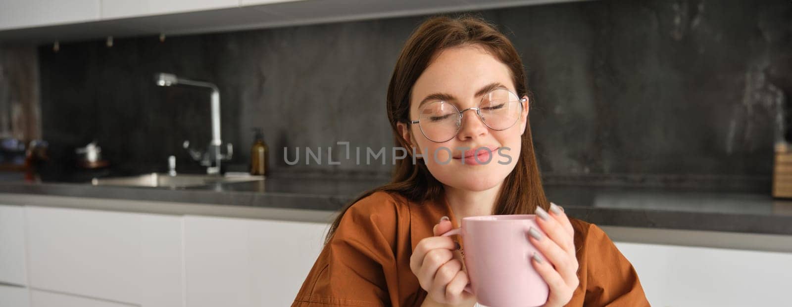 Portrait of beautiful woman in glasses, enjoying cosy autumn day at home, holding cup of coffee and smells great flavour and aroma, smiling pleased.