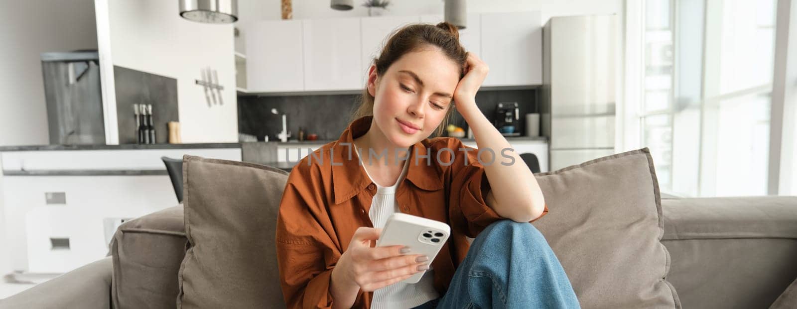 Portrait of young modern woman reading on mobile phone, scrolling social media app on smartphone, sitting on couch in living room in casual clothes.