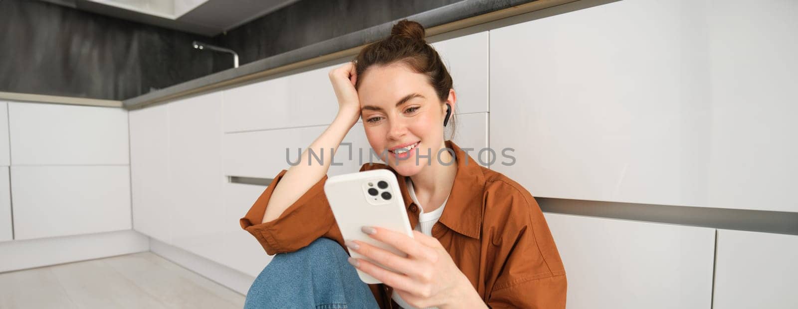 Portrait of happy young beautiful woman, listening music in wireless earphones, using smartphone, watching videos on mobile phone with headphones on.