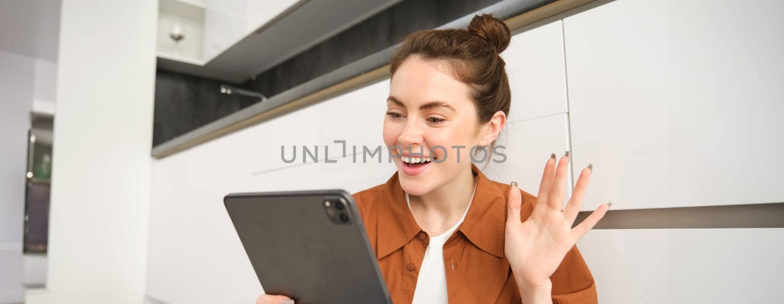 Portrait of friendly smiling woman video chats, connects to online conversation on digital tablet, says hello at camera.