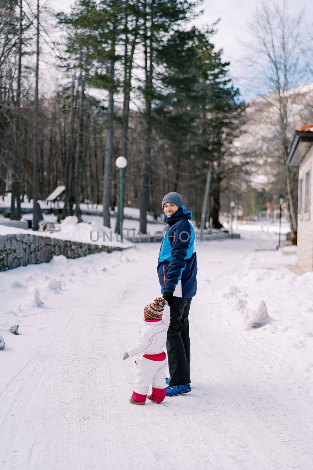 Smiling dad with a small child are standing on a snowy road near the house, holding hands. Side view by Nadtochiy