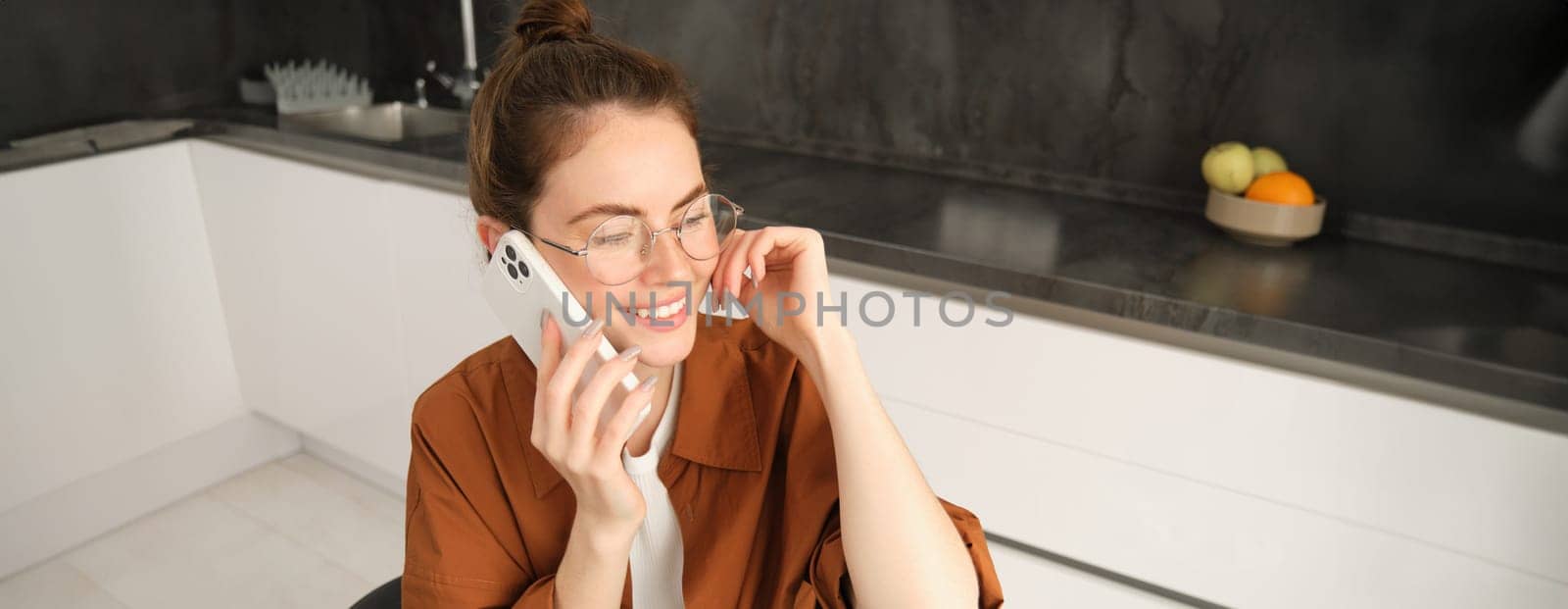 Modern young woman sitting in kitchen with smartphone, talking on mobile phone, has friendly conversation, calling someone.