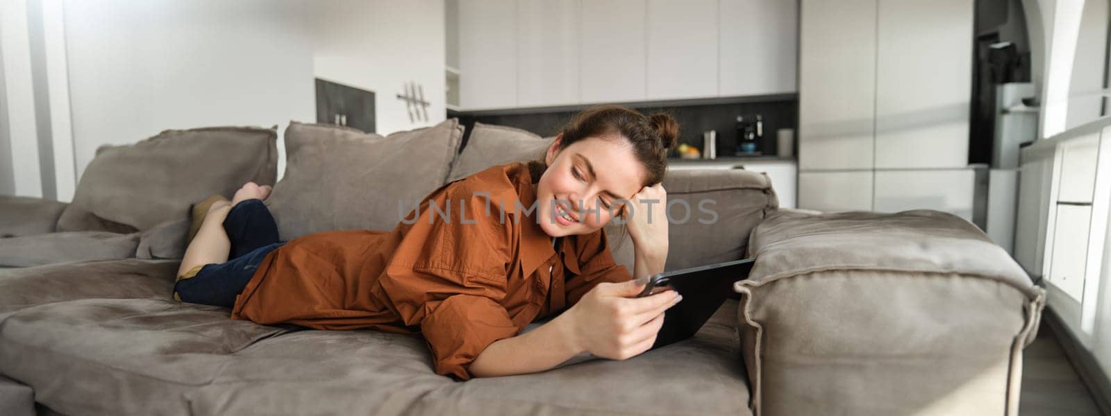 Portrait of beautiful young woman resting at home on couch, lying on sofa with digital tablet, reading or watching videos on gadget.