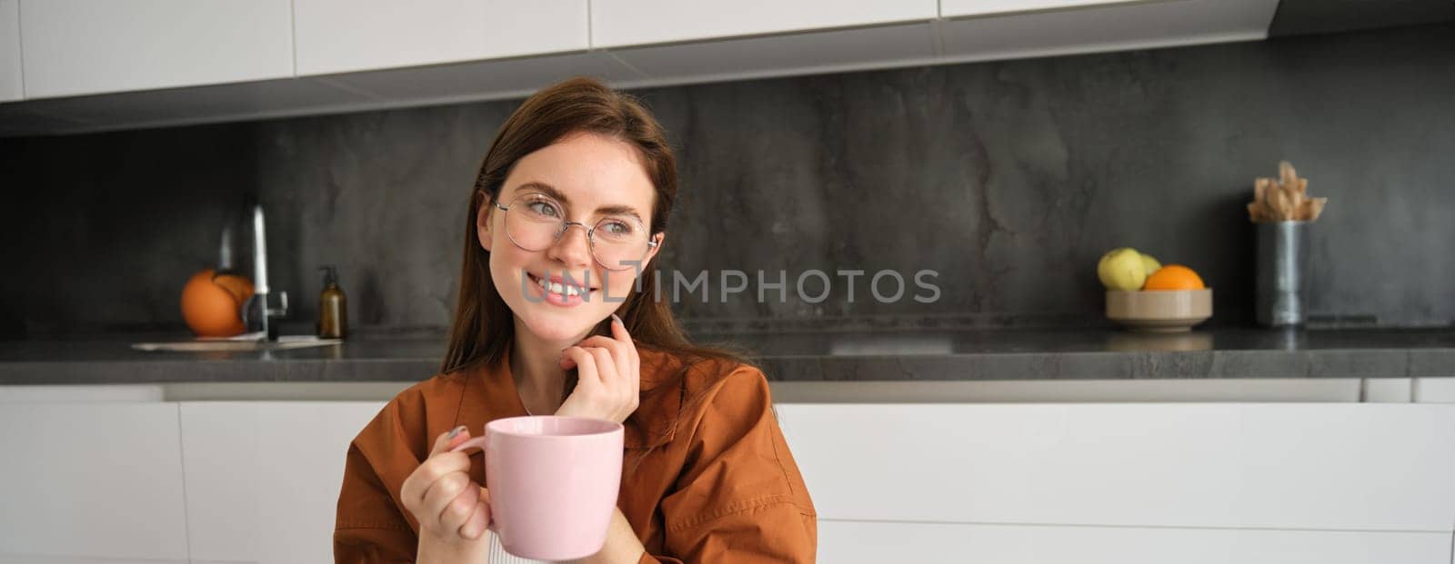 Portrait of young brunette woman in kitchen, resting indoors, smiling and drinking coffee, enjoys tasty hot tea.