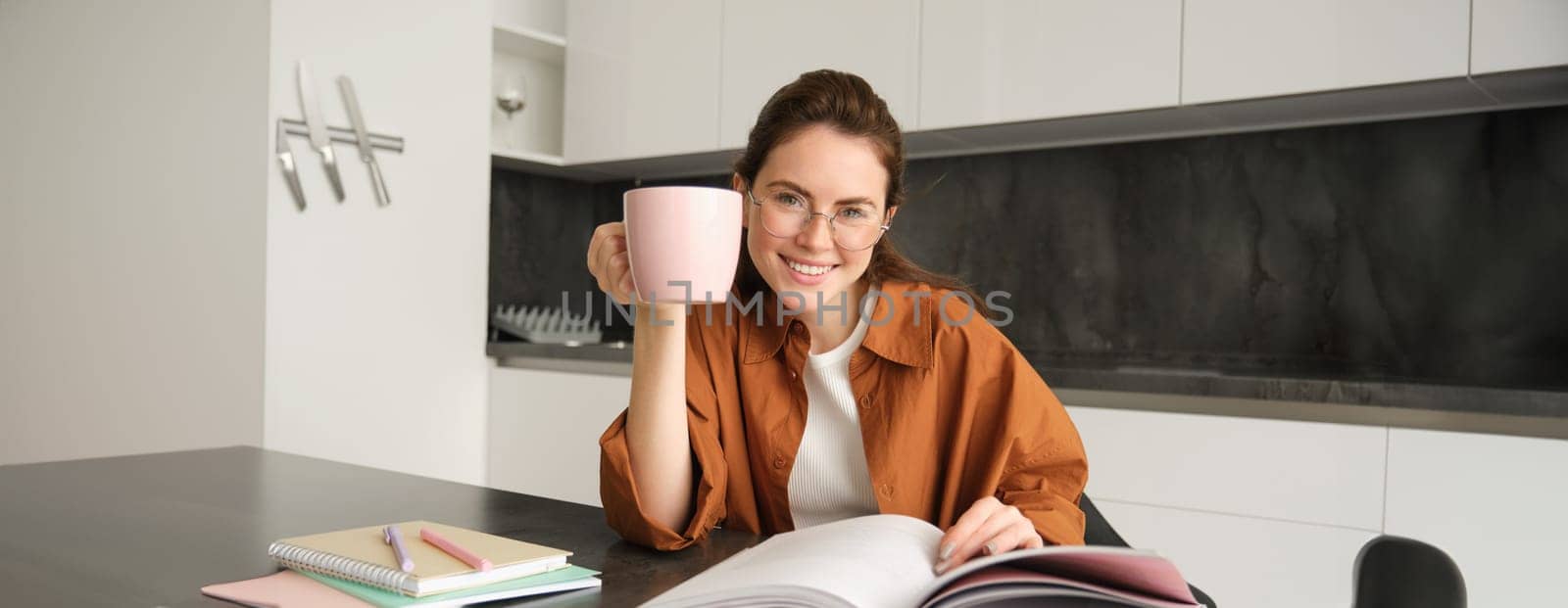 Distance learning. Young woman at home, student studying at home and drinking tea, reading her work book, revising for exam by Benzoix