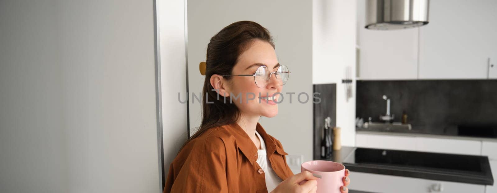 Portrait of happy young woman at home, holding cup of tea, leaning on wall, smiling, drinking coffee from pink mug.