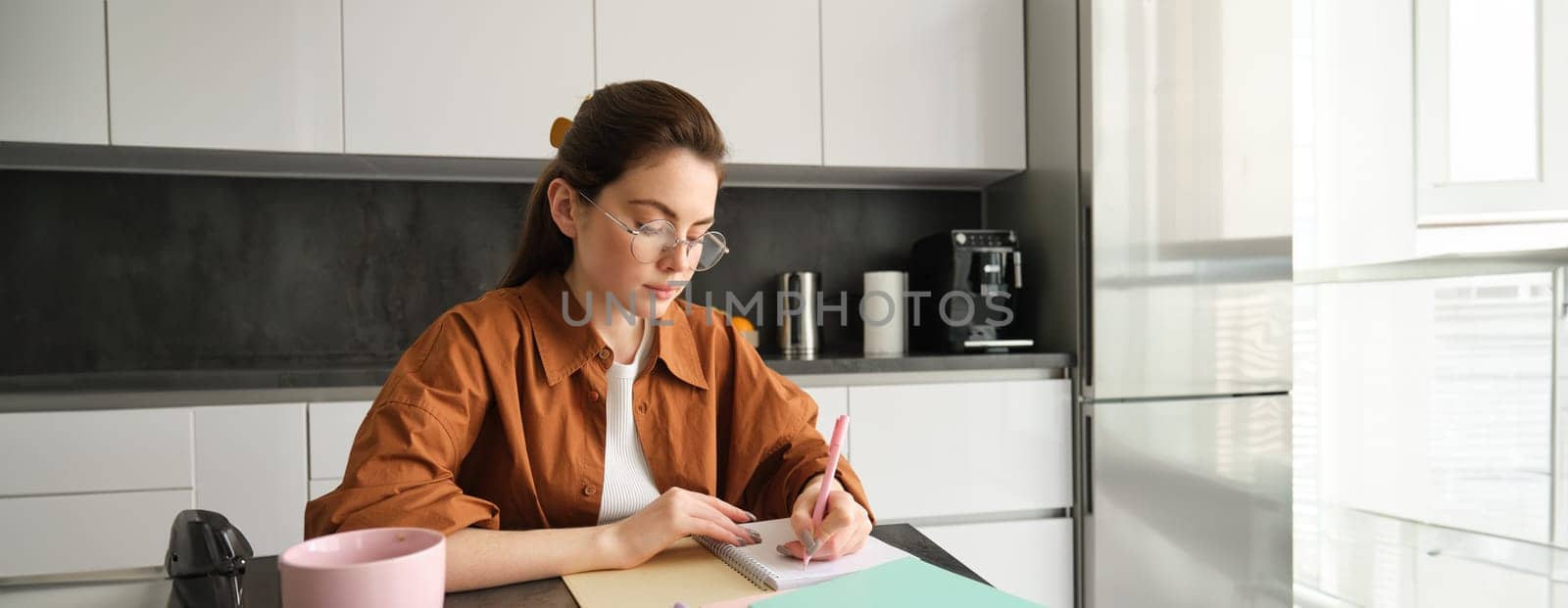 Portrait of woman looking concentrated, writing down notes, doing homework in kitchen, drinking coffee, studying at home by Benzoix
