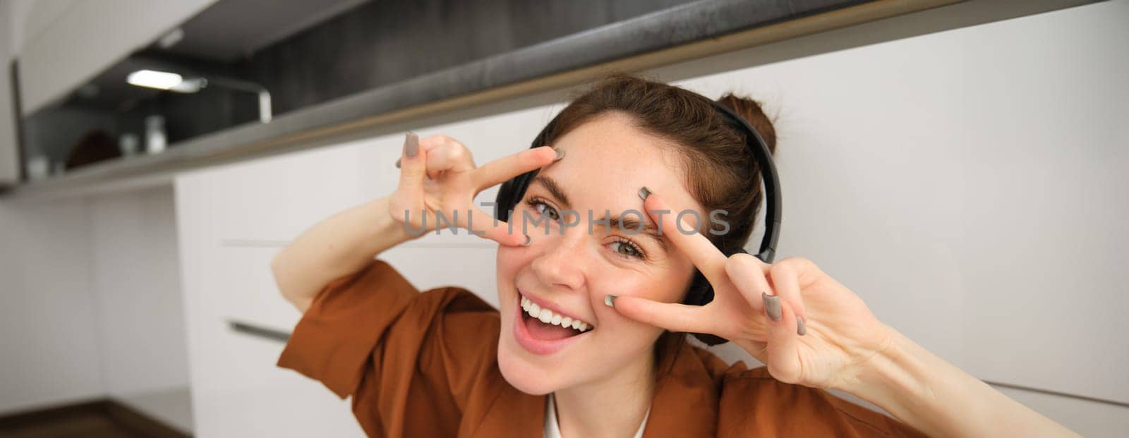 Happy female model listens music, posing in wireless headphones, smiling and laughing, showing v-sign.