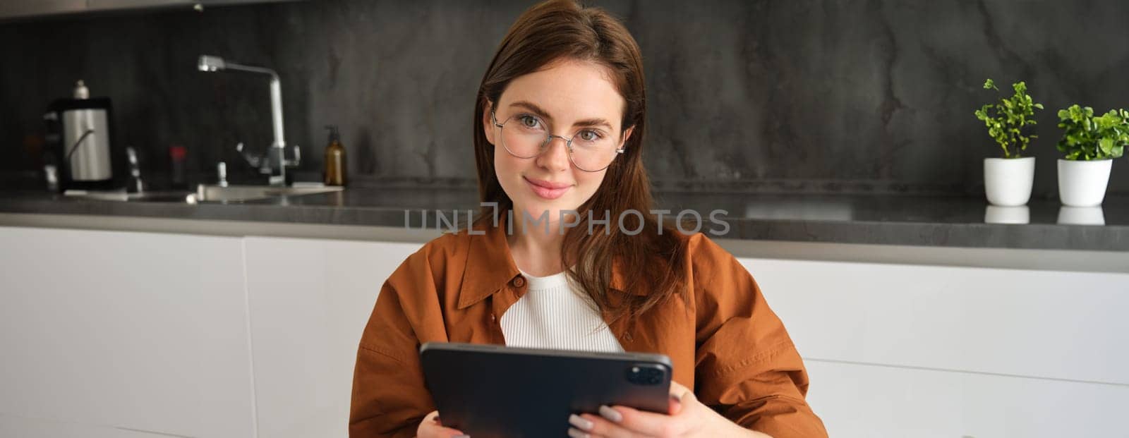 Portrait of brunette woman in glasses, sitting in kitchen at home, holding digital tablet, reading on gadget.