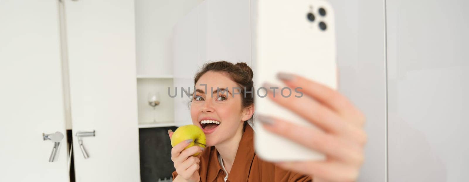 Beautiful young woman, lifestyle blogger records video of herself, takes selfie while biting an apple, sitting on kitchen counter, using smartphone.