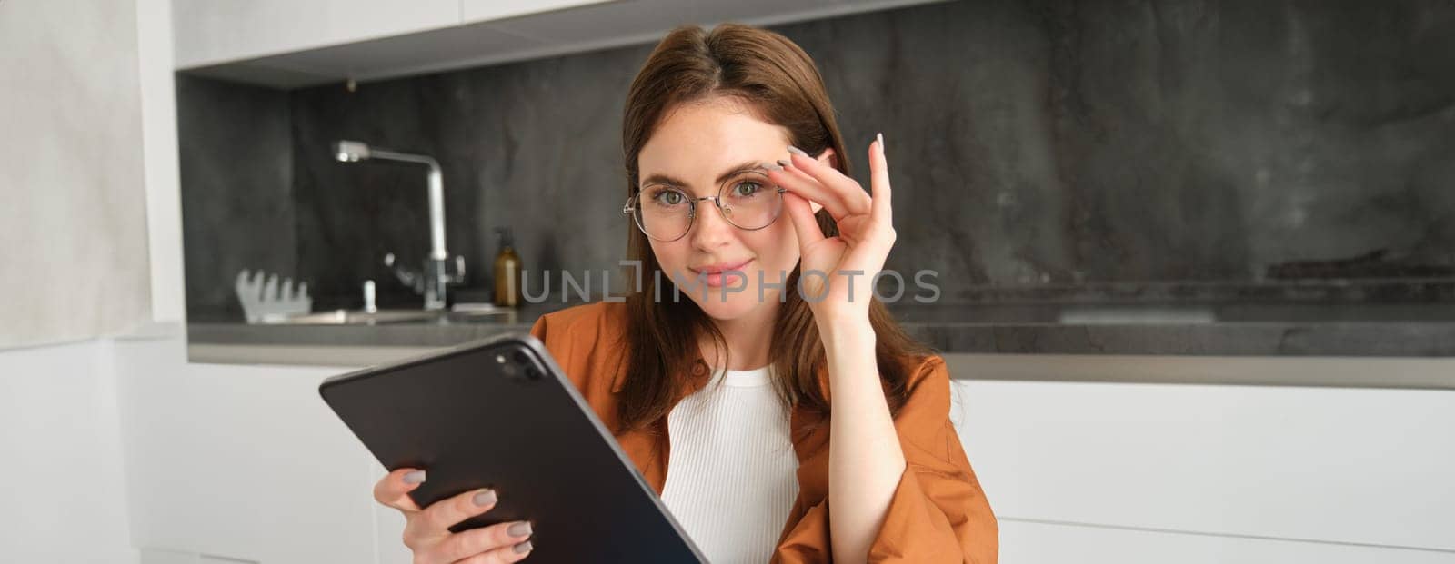 Portrait of beautiful young female model in glasses, holding digital tablet, wearing glasses, reading e-book, posing in kitchen at home.