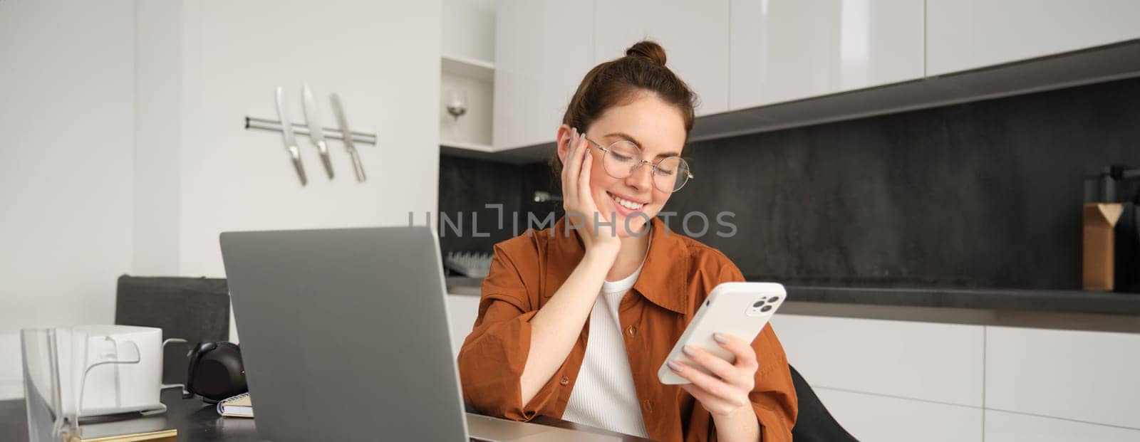 Portrait of young woman freelancer, working from home, saleswoman doing trading from her kitchen, set up workplace in flat, sitting near laptop, using smartphone.