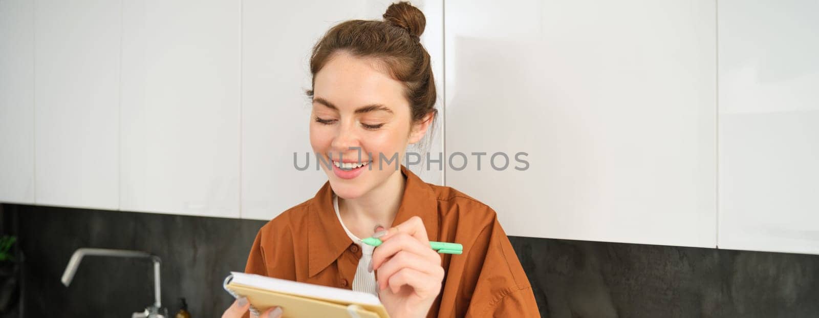 Portrait of smiling young woman with notebook, writing, making notes in planner, sitting at home in the kitchen.