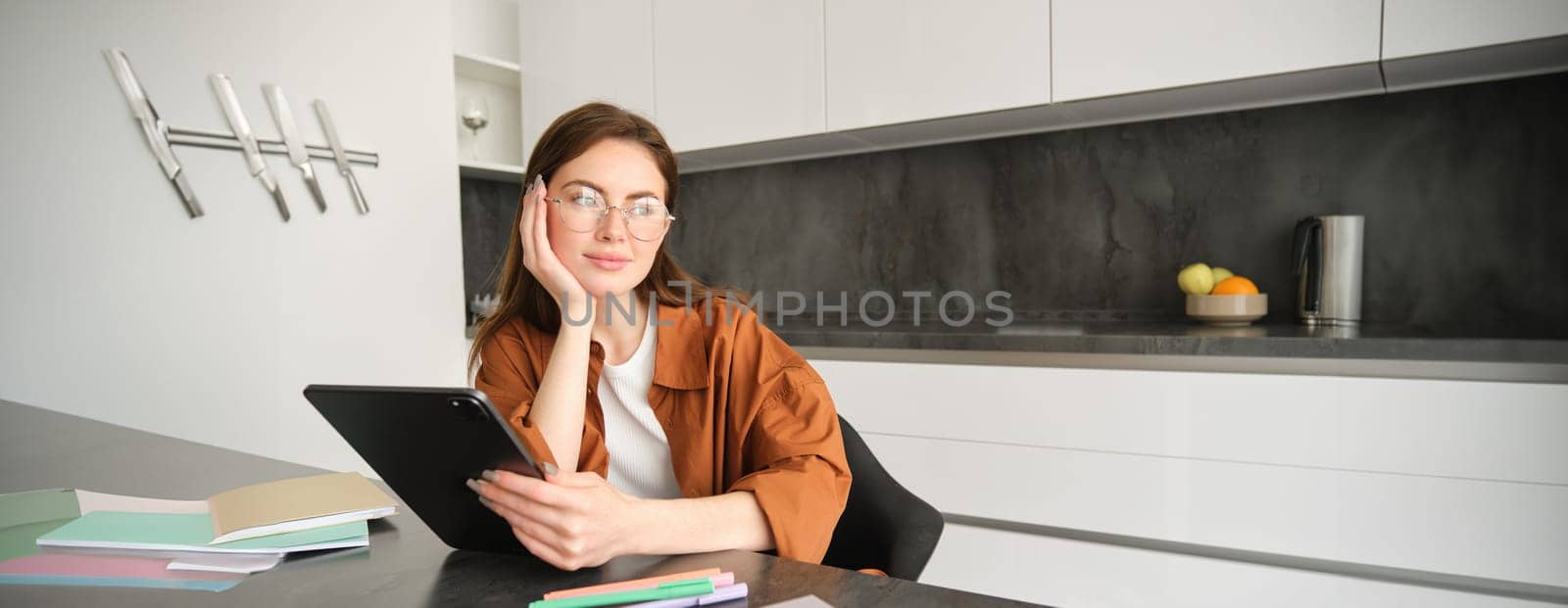 Portrait of young student, woman studying at home, working remotely, setup workplace in her kitchen, sitting on chair with digital tablet, reading in glasses by Benzoix