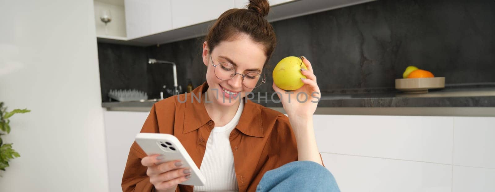 Portrait of stylish young woman in glasses, sitting in the kitchen with mobile phone and eating apple, checking social media, using smartphone app.