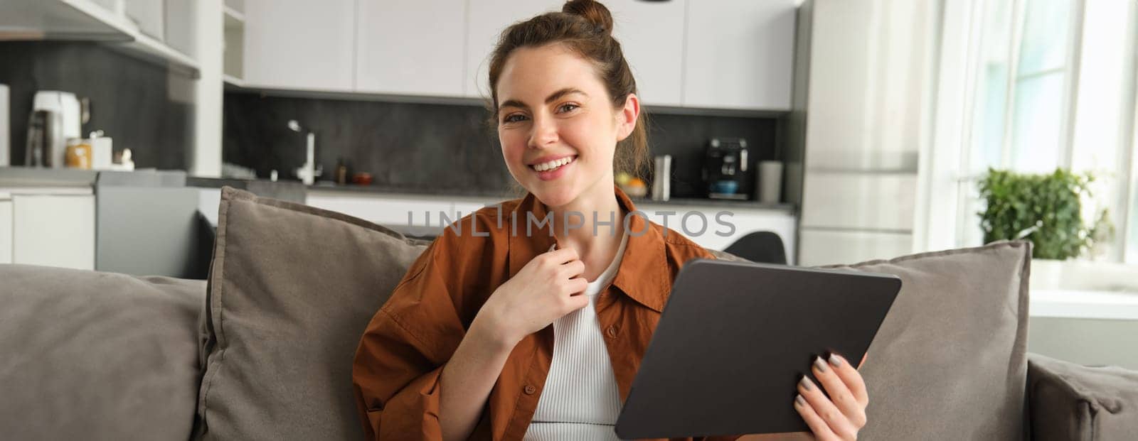 Cute young brunette woman holding digital tablet in hand, sitting on couch, smiling and laughing at camera.