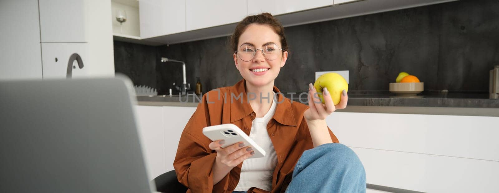 Image of young modern woman, relaxing at home, sitting on chair and using smartphone, eating green apple and smiling by Benzoix