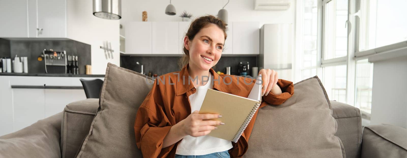 Smiling young brunette woman, sits on sofa in living room, holds notebook, reads her notes, studies for exam, student does her homework at home.