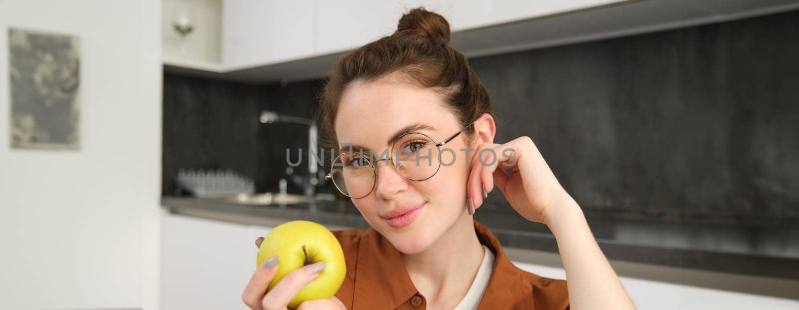 Beautiful young woman in glasses, sitting on chair in the kitchen, eating green apple and smiling.
