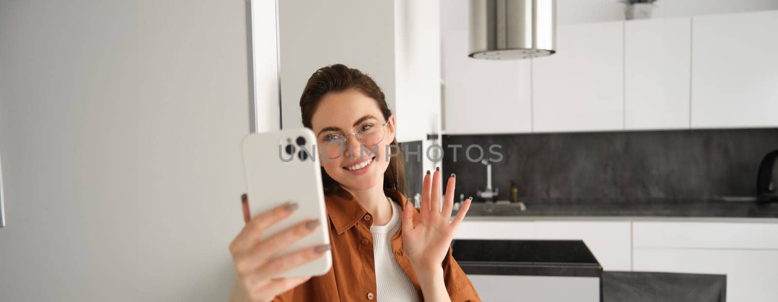 Portrait of smiling young woman standing with mobile phone at home, waving hand at smartphone, connects to online chat, talking to someone.