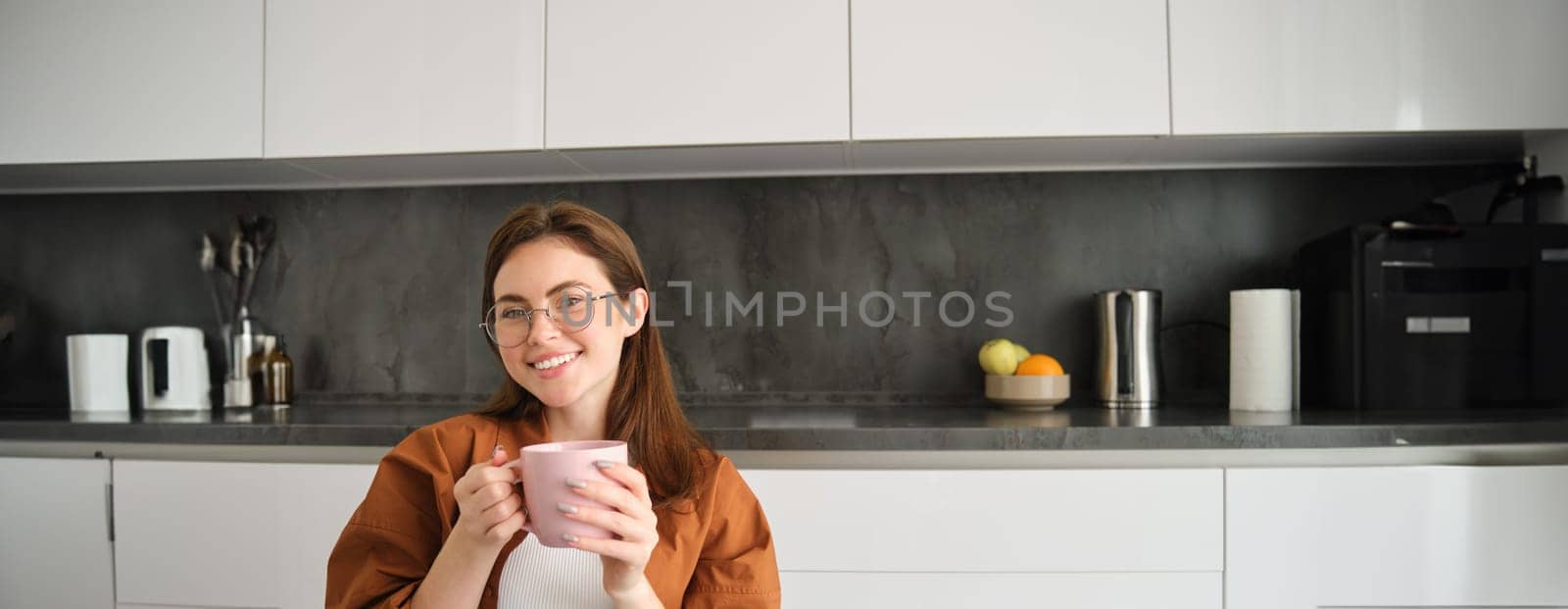 Portrait of happy young woman in glasses, sitting at home in kitchen, holding cup of coffee, drinking favourite tea, relaxing indoors on cold autumn day.