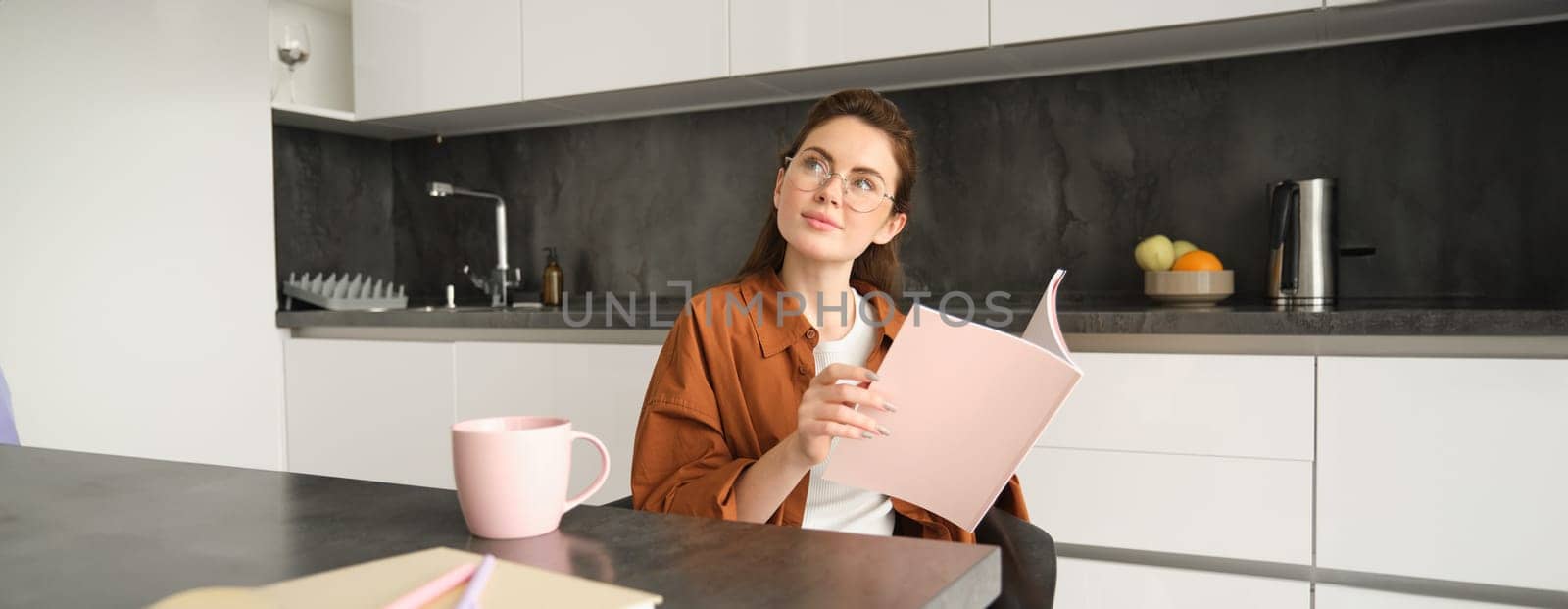 Portrait of young woman working from home, self-employed entrepreneur reading her documents, sitting at home in kitchen, wearing glasses.