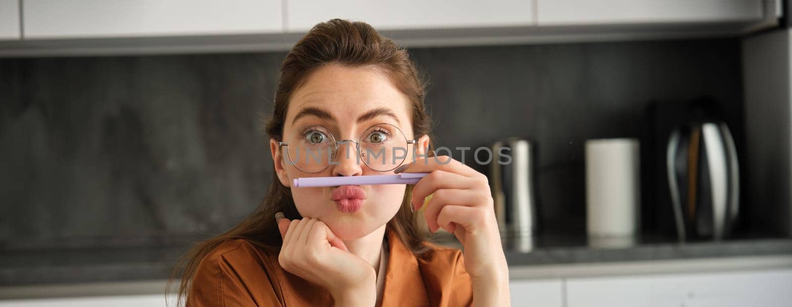 Close up portrait of funny, silly young woman, playing with pen, holding pencil on top of her lip and grimacing, sitting in kitchen by Benzoix