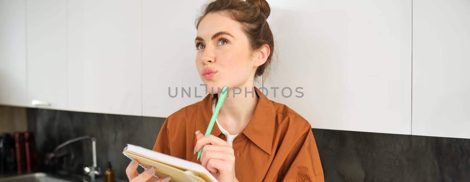 Close up of thoughtful young woman with notebook, looking up with thinking face, writing down her ideas or thoughts in planner, makes list of groceries by Benzoix