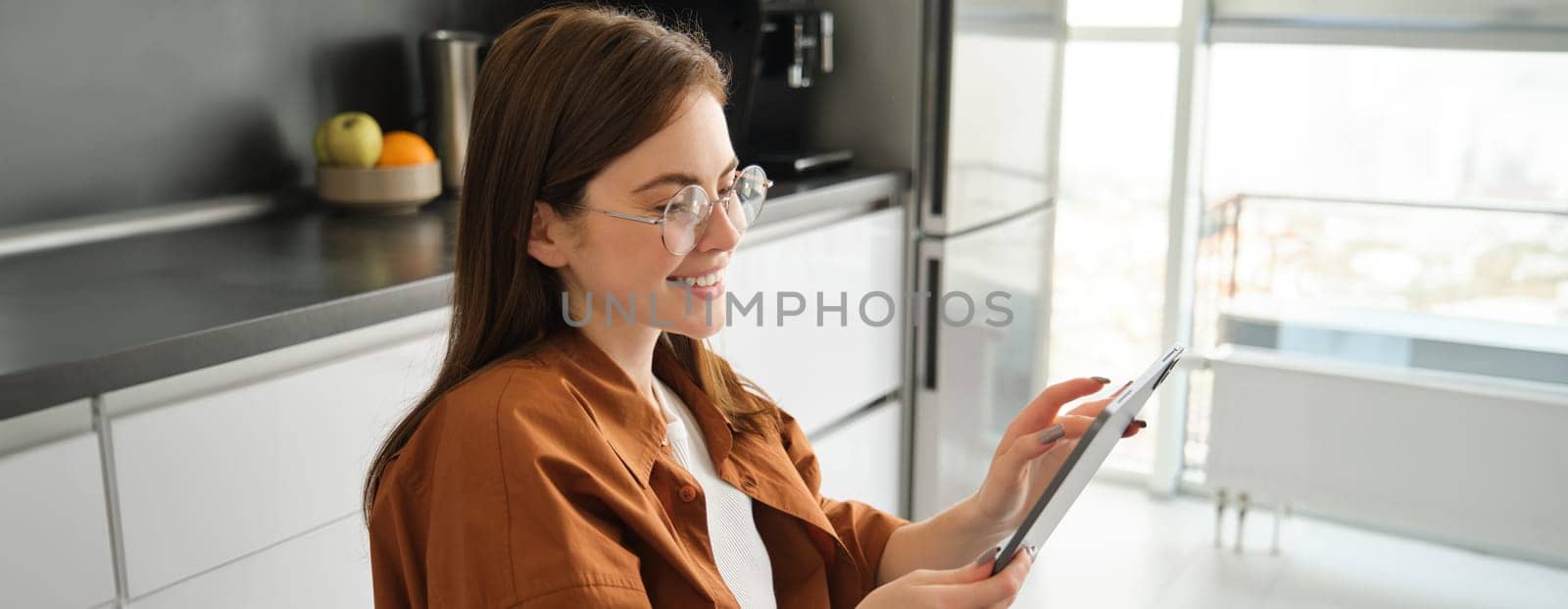 Side view of young woman in glasses, beautiful girl reading on digital tablet, messaging, online shopping with gadget, smiling, sitting in kitchen at home.